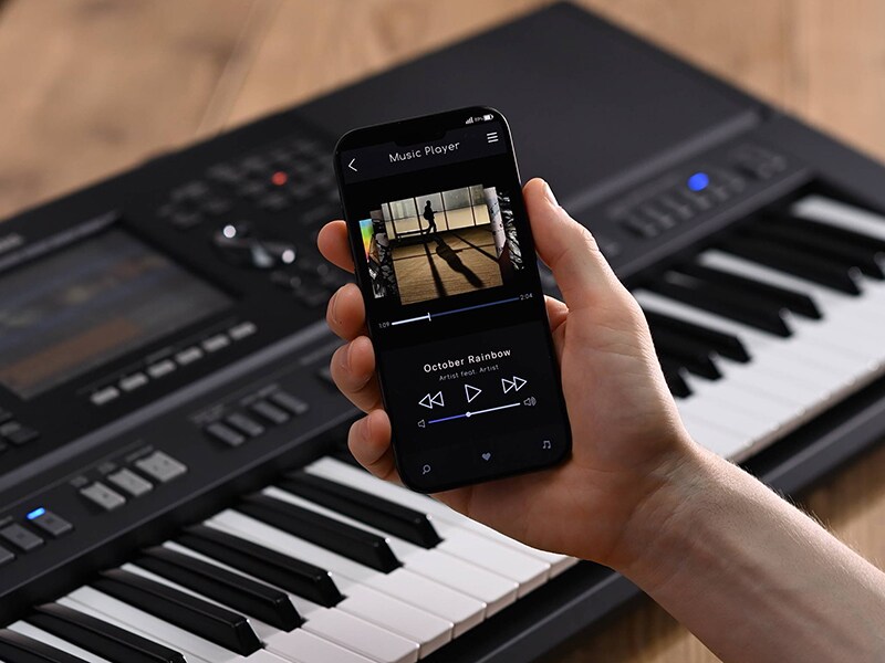 Yamaha Arranger Workstation PSR‑SX920 y una mano sosteniendo un smartphone con la pantalla de un reproductor de música