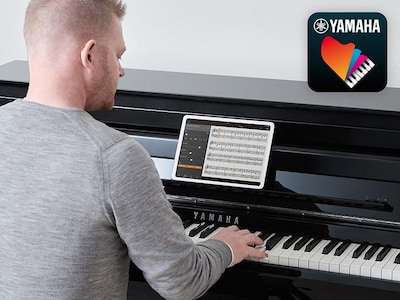 A person playing the piano by placing the smart device on a music stand and displaying sheet music on the Yamaha Smart Pianist app screen, and the Smart Pianist app icon
