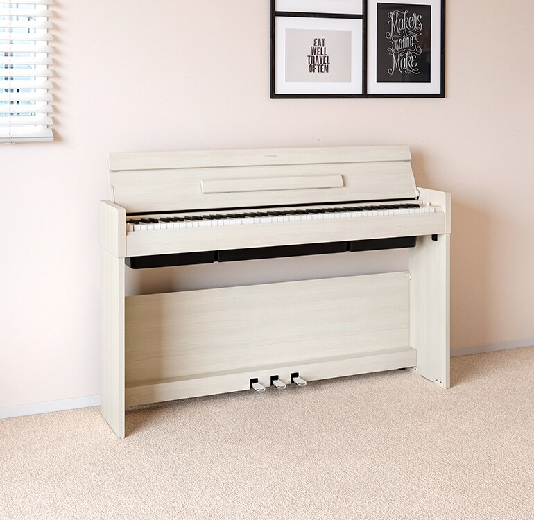 Un salón con un piano ARIUS en abedul blanco situado junto a unas estanterías de pared con lámparas y un difusor, integrado de forma armoniosa en la decoración.